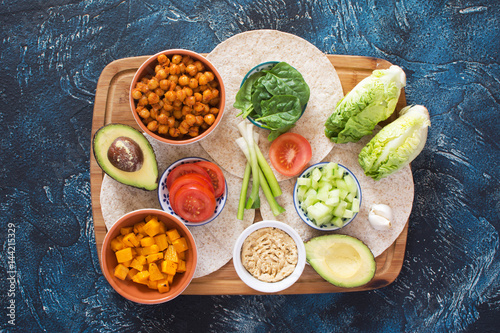 Ingredients for vegetarian tortillas on the wooden board chickpeas, butternut squash, spring onion, cucumber, avocado on the dark blue table, top view, copy space for text