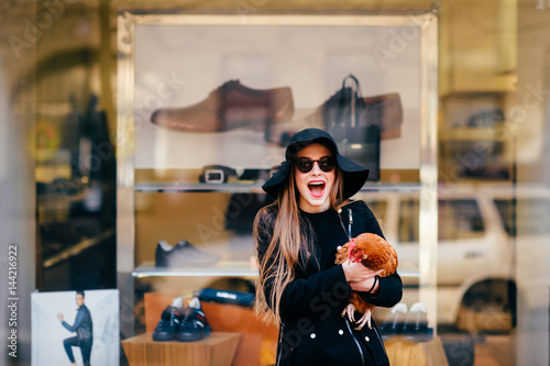 Portrait of beautiful and lovely model in fashion clothes  black sunglasses and elegant hat standing in front of boutique showcase with chicken in hands and laughing. Unusual pets concept. Expressive.