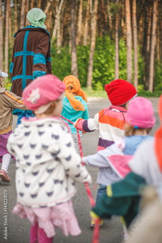 Group of little kids with a kindergartener walking to the forest and hold a colorful rope. Middle spring green pine forest hike in Waldorf kindergarten.