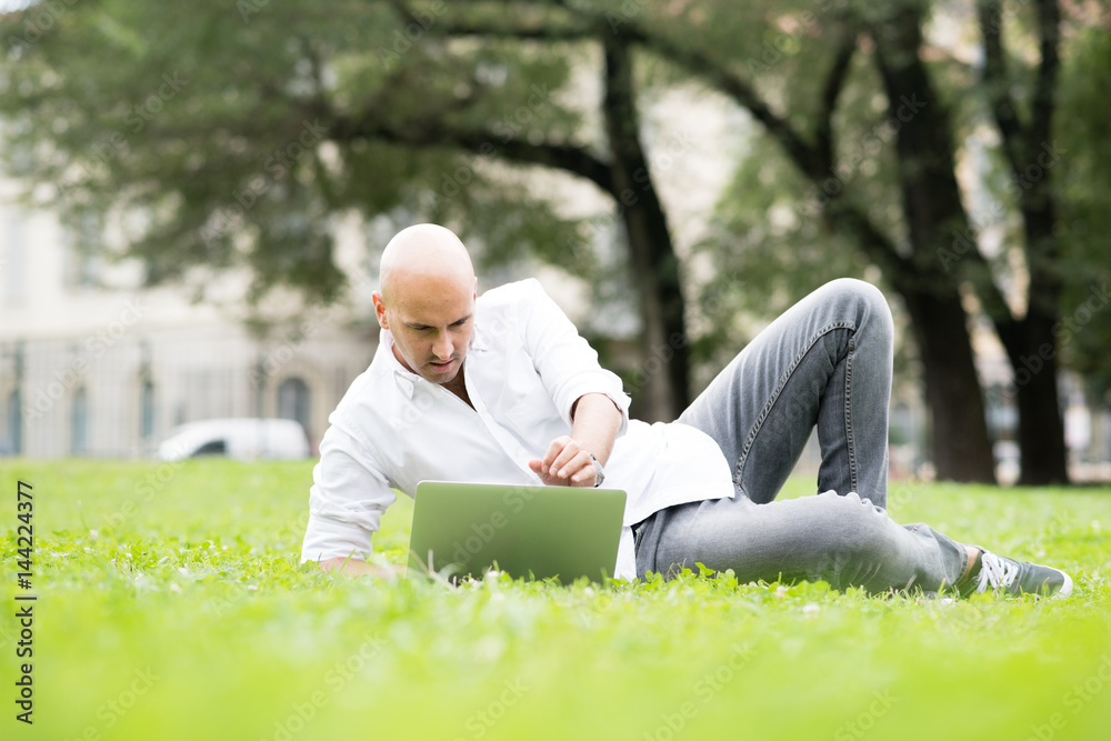 Fototapeta premium Young contemporary businessman remote working sitting outdoor in a city park using computer - portability, small business, networking concept
