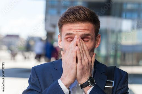 Young man in suit manager sneezes on the street covering his face with his hands. Allergic reaction in the spring on the street.