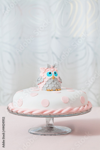 Birthday cake covered with fondant displayed on the pink cloth and glass tray  decorated with pink dots and an grey and pink fondant owl with blue eyes sitting on top