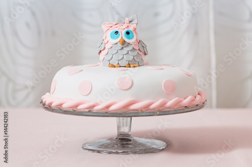 Birthday cake covered with fondant displayed on the pink cloth and glass tray; decorated with pink dots and an grey and pink fondant owl with blue eyes sitting on top