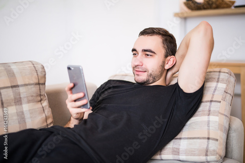 Beautiful young man relaxing on sofa and looking at his smart phone at home