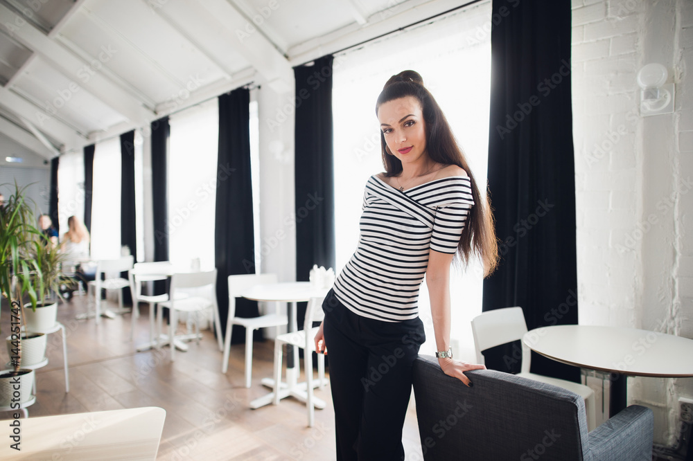 Attractive female owner of a modern cafe standing near wooden table with huge windows on the background. Beautiful woman leaning against a chair and looking at the camera. Attractive female owner of a modern cafe standing near wooden table with huge windows on the background. Beautiful woman leaning against a chair and looking at the camera.