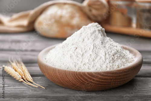 Flat bowl of white flour and wheat ears on wooden table