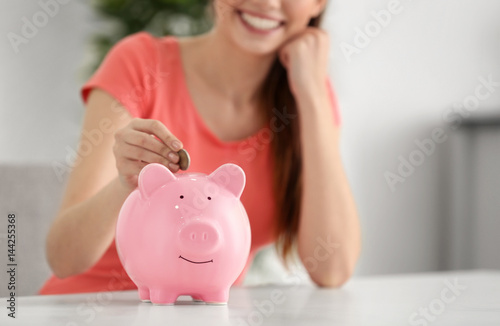 Young woman putting money into piggy bank at home