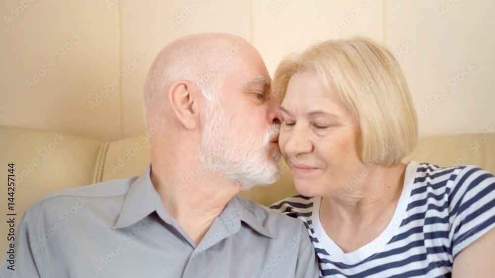Cheerful good-looking senior couple sitting on sofa at home. Kissing smiling having good time. Cheerful good-looking senior couple sitting on sofa at home. Kissing smiling having good time.