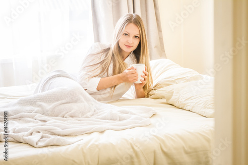 indoor portrait of young pretty happy woman with a cup of tea or coffee in bed, morning at home