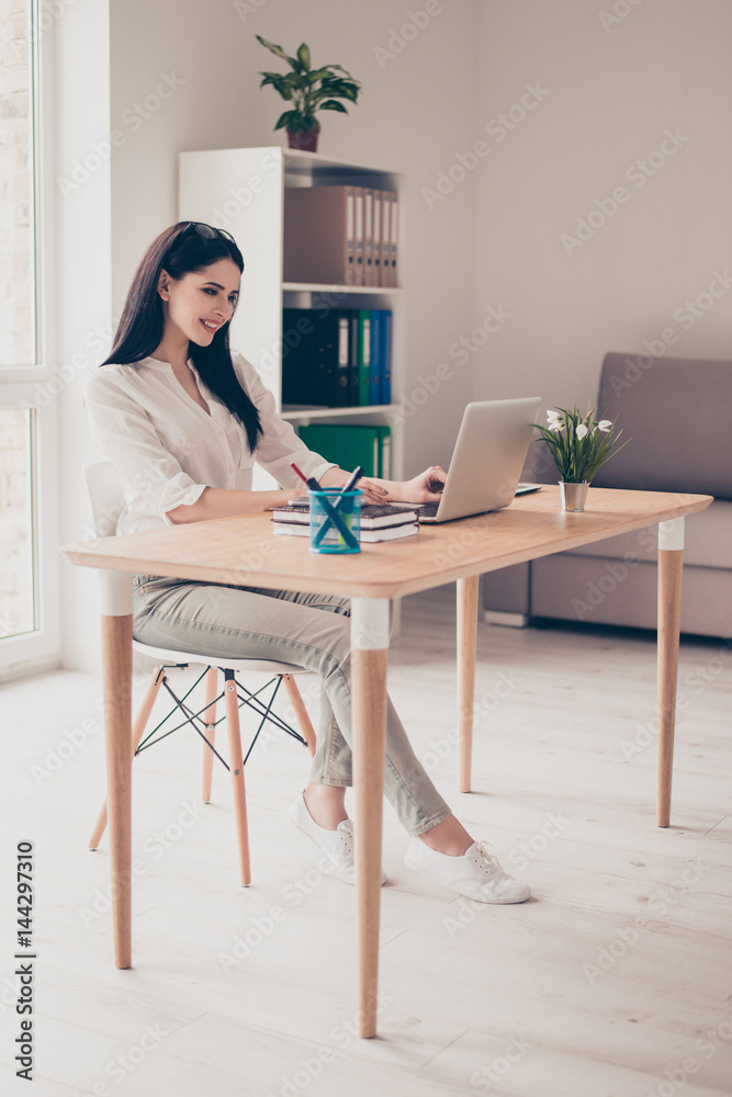 Cute young  woman sits at the desk in the office, works with  laptop and looks at the screen