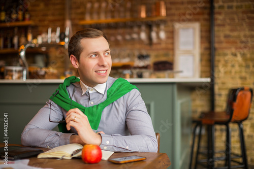 Happy businessman sits at the table with working things and keeps glasses in his hand in time of a break, in a cafe