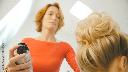 Professional hairdresser, stylist doing hairstyle for young pretty woman and using hair spray in white make up room. Beauty and haircare concept