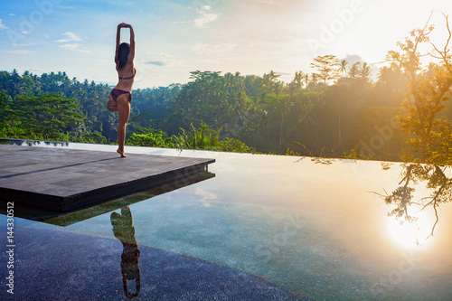 Good morning with yoga meditating on sunrise background. Active woman in bikini practicing on villa poolside to keep fit and health. Woman fitness training, sport activity on summer family holiday.