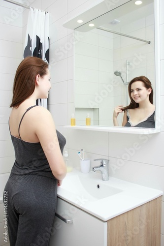 morning and people concept - young woman looking to mirror at home bathroom