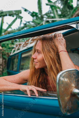 Red hair surf girl near blue vintage mini van with palm trees in the background in grey swimsuit, Indonesia, Bali