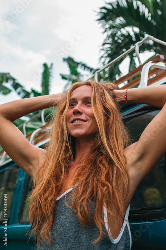 Red hair surf girl near blue vintage mini van with palm trees in the background in grey swimsuit, Indonesia, Bali