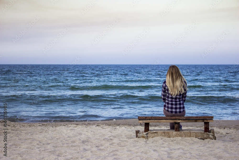 Lonely girl sitting on a bench on the background of the sea , sunset landscape .