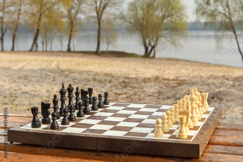 Chess board with chess pieces on bench with river embankment background. Chess game