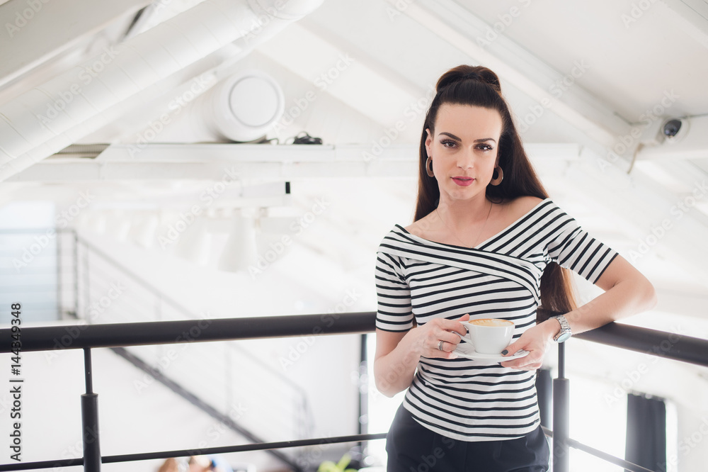 Beautiful woman with a cup of aromatic coffee standing above main room and looking at the camera.