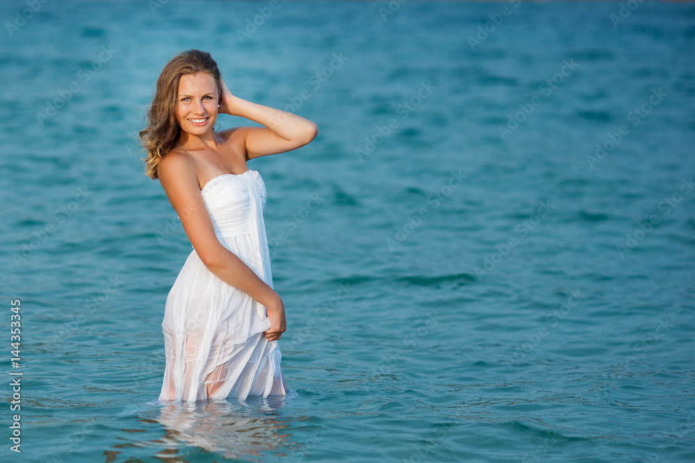 Beautiful woman standing in the sea in white dress. Pretty girl got wet her dress in the sea water. Attractive woman smiling at camera. Beautiful woman standing in the sea in white dress. Pretty girl got wet her dress in the sea water. Attractive woman smiling at camera.