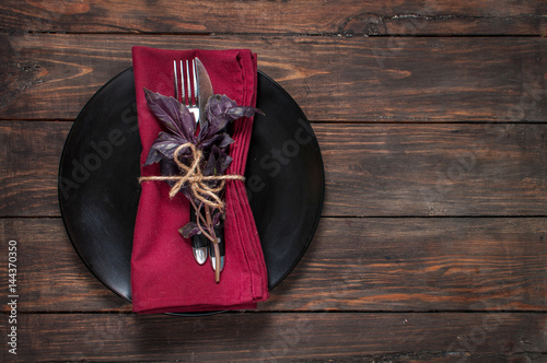 Black plate with fork,knife, napkin and basil on wooden table