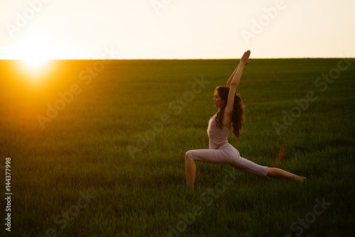 Young woman doing complex Yoga exercise on green lawn at sunset