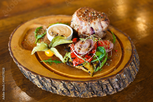 The close-up side view of the grilled meat and salad consisted of the onions, chilli, bazilik placed near the cup with sauce on the wooden board. photo