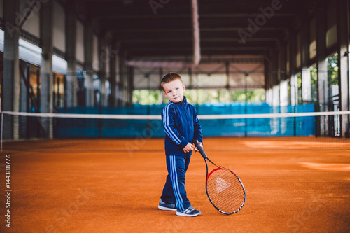 little boy on the yellow, sandy tennis court stands with racket in blue tracksuit © Elizaveta