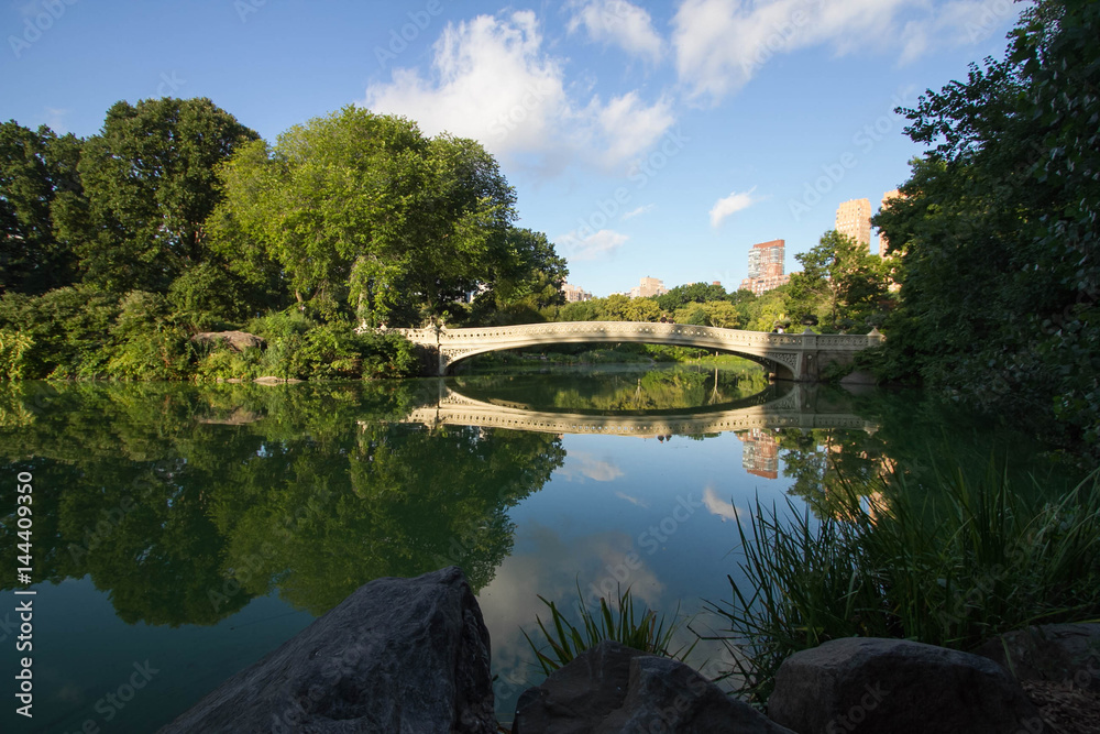Bow bridge and big trees reflect in lake with cloudy sky and rocks under the shade at Central Park