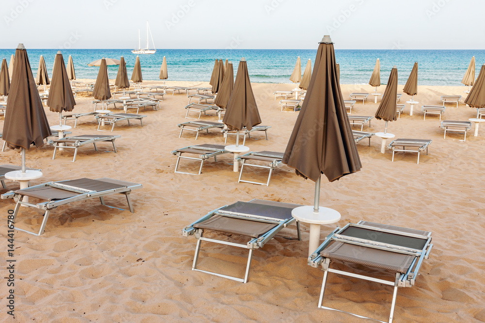 Empty beach, loungers and closed parasols with ocean and lonely yacht in the background