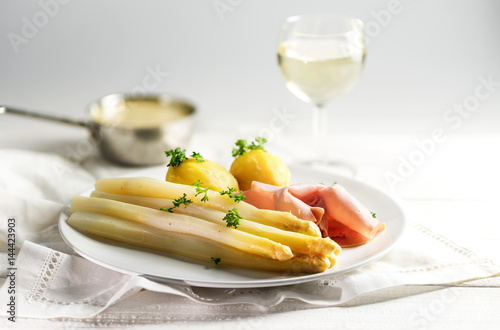 Juicy white asparagus dish with potatoes and ham on a white plate, sauce hollandaise and wine blurred in the bright background, copy space