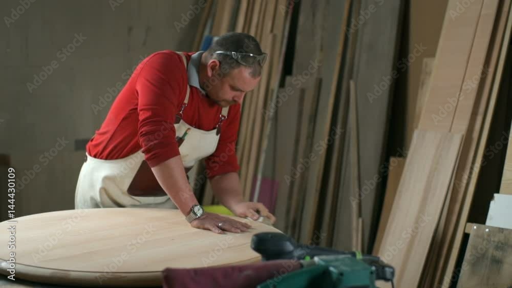 Carpenter leans on a table and polishes it with an abrasive block in workshop Carpenter leans on a table and polishes it with an abrasive block in workshop