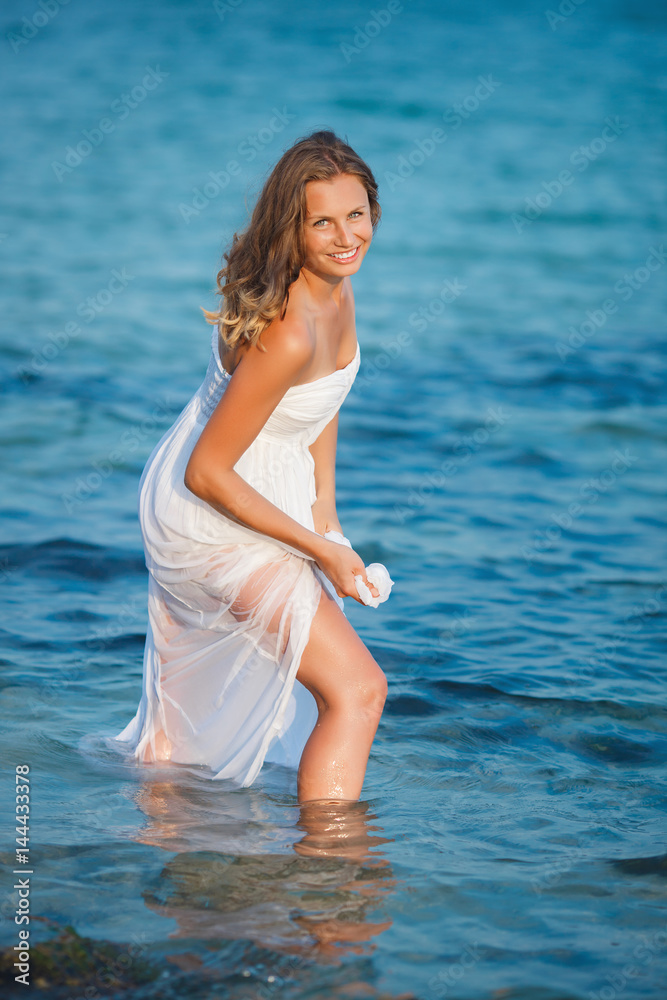 Beautiful woman standing in the sea in white dress. Pretty girl got wet her dress in the sea water. Attractive woman smiling at camera. Beautiful woman standing in the sea in white dress. Pretty girl got wet her dress in the sea water. Attractive woman smiling at camera.