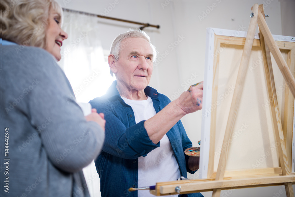 Happy senior couple painting picture on easel at home Happy senior couple painting picture on easel at home