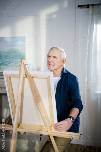 Pensive senior man painting picture on easel at home