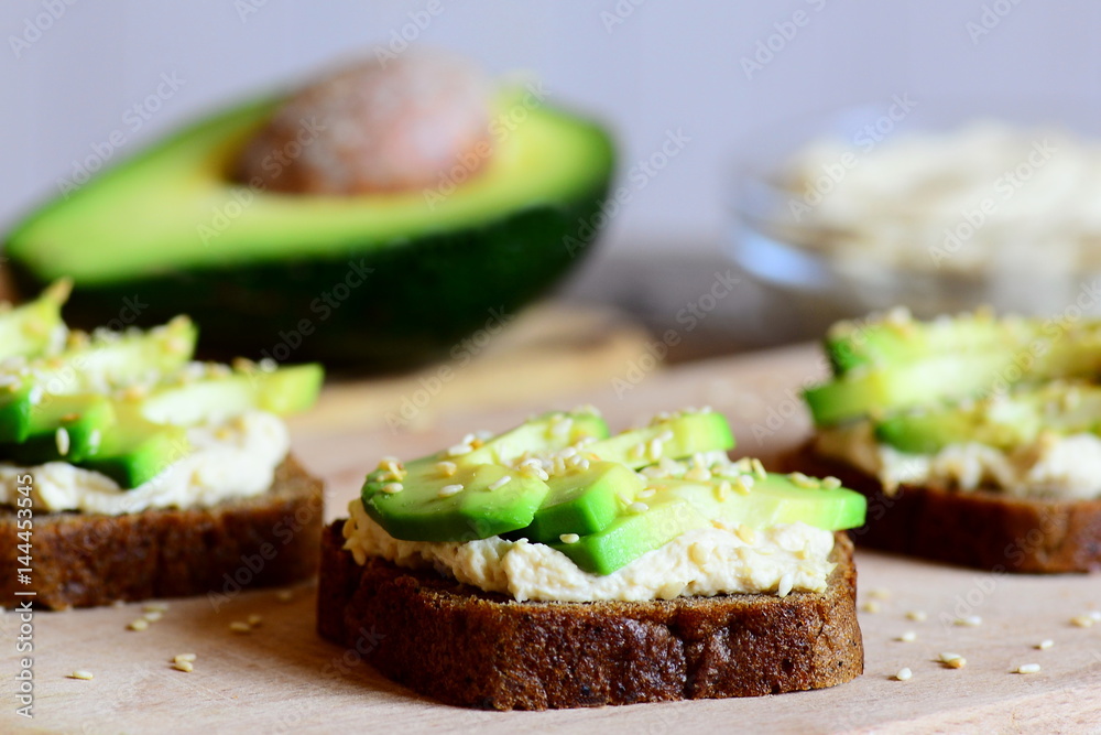 Hearty chickpeas hummus and avocado sandwiches on a wooden board, avocado half, hummus in a glass bowl. Veggie sandwiches cooked with rye bread, avocado slices, chickpeas hummus and fried sesame seeds