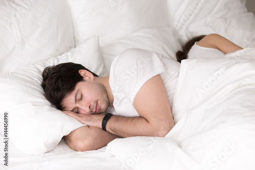 Young guy and lady asleep together, having good dreams, healthy sleep, couple in bed taking a nap back to back. Sleeping man wearing smart wristband tracker for sleep tracking and getting up on time