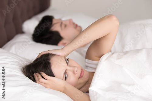 Annoyed upset wife trying to sleep with snoring husband asleep next to her, young girl waking up with headache, can not sleep, having insomnia, feeling bad in the morning after sleepless night