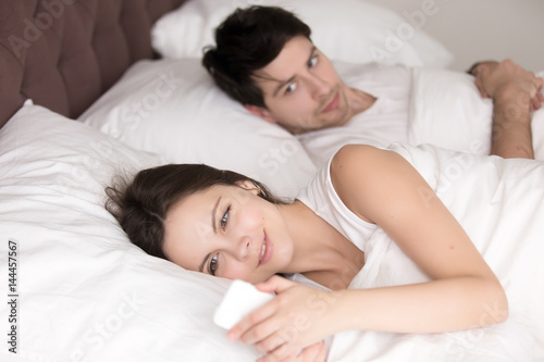 Couple in bed, happy smiling woman turned her back to man, reading message on phone from her lover, worried boyfriend lying next to her, trying to peek at screen. Cheating and infidelity concept