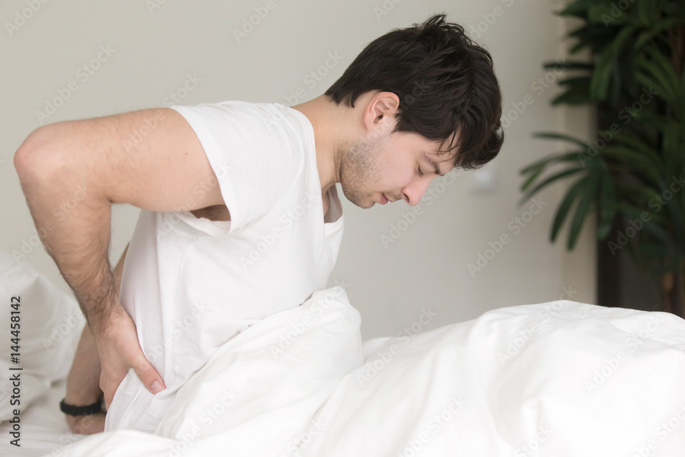 Young guy wearing white t-shirt sitting indoor on the bed feeling sudden ache in his back, suffering from backpain, need to make doctor s appointment. Healthcare and treatment concept Young guy wearing white t-shirt sitting indoor on the bed feeling sudden ache in his back, suffering from backpain, need to make doctor s appointment. Healthcare and treatment concept
