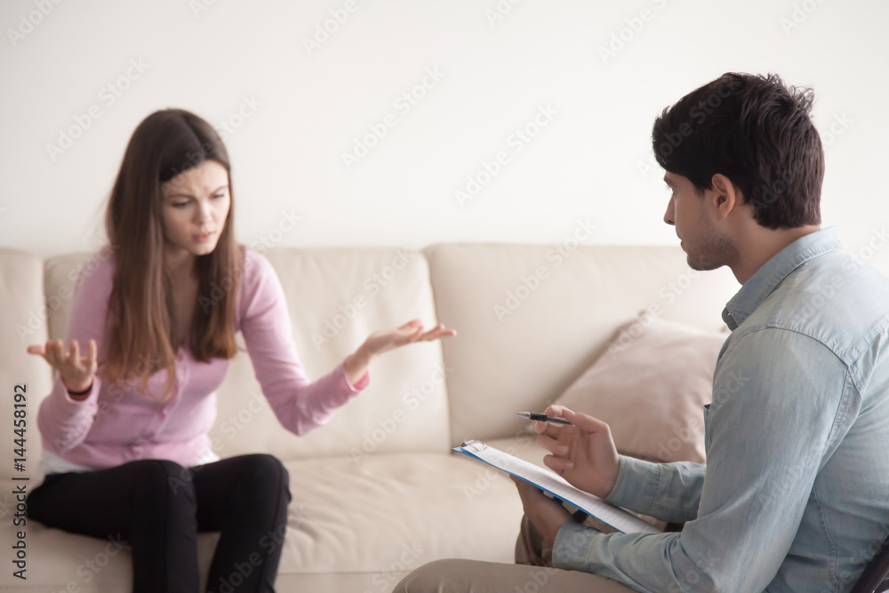 Young lady at psychologists office. Frustrated sad girl, having problems at work or in relationships, sitting on sofa telling about her problems and describing her feelings, male doctor making notes Young lady at psychologists office. Frustrated sad girl, having problems at work or in relationships, sitting on sofa telling about her problems and describing her feelings, male doctor making notes