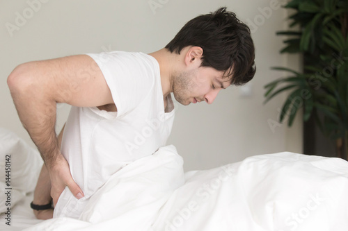 Young guy wearing white t-shirt sitting indoor on the bed feeling sudden ache in his back, suffering from backpain, need to make doctor s appointment. Healthcare and treatment concept