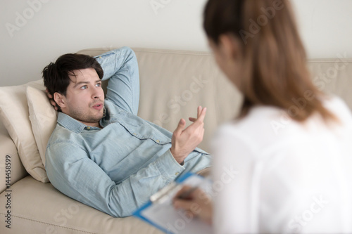 Female psychologist working with young man lying comfortably on couch and talking about his problems. Guy on reception at psychotherapist office, trying to relax and describe his feelings