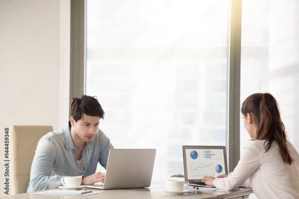 Business partners working together, using two separate laptops, drinking coffee and looking at screen, man and woman sitting opposite at the same desk in office near big window. Copy space Business partners working together, using two separate laptops, drinking coffee and looking at screen, man and woman sitting opposite at the same desk in office near big window. Copy space