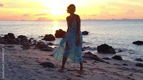 Happy caucasian woman in a long dress walks on the beach on a colourful sunset background