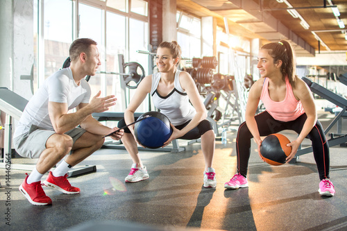 Two sporty women doing exercises with fitness balls with assistance of their personal trainer in gym.