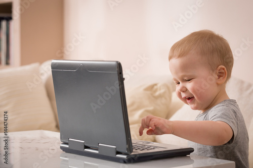 Cute little toddler boy sitting on the sofa with his laptop and looking at the window. Learning with computer. Early development. Child and computer.
