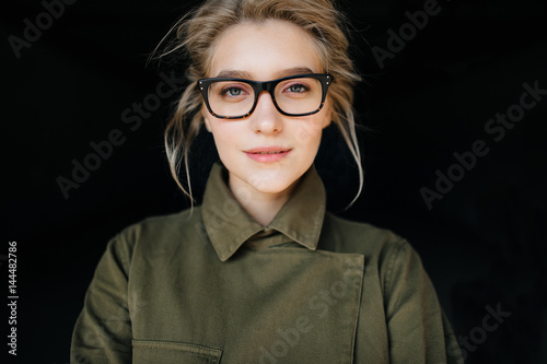Close up face portrait of beautiful, cute attractive young model blonde fashion student girl in coat, glasses and curly hair looking at camera. Isolated on black. Tenderness, femininity and charming.
