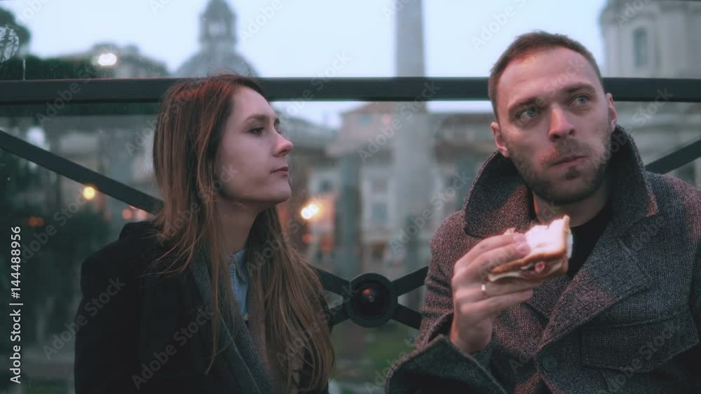 Young stylish couple sitting at the city centre and eating sandwiches in evening. Looks aside together. Young stylish couple sitting at the city centre and eating sandwiches in evening. Looks aside together.