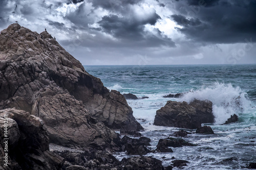 Foggy view of Pebble Beach California coast with storm clouds and rough seas causing waves to crash on rocks. The beach is shaped by erosion and climate change.